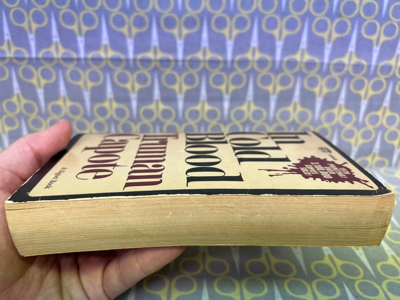 May include: A vintage paperback copy of Truman Capote's In Cold Blood. The book cover features the title in bold black font with a red blood splatter graphic.