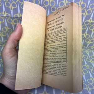 May include: A close-up of a book's back cover with a handwritten inscription and text praising Truman Capote's novel "In Cold Blood". The text highlights the book's status as a bestseller and a great modern crime story.