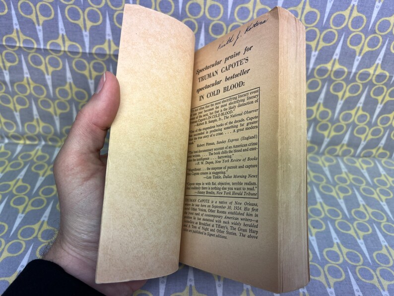 May include: A close-up of a book's back cover with a handwritten inscription and text praising Truman Capote's novel "In Cold Blood". The text highlights the book's status as a bestseller and a great modern crime story.
