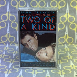 May include: A cassette tape with a blue and red title that reads "Two of a Kind" featuring John Travolta and Olivia Newton-John. The cassette tape is in a clear plastic case. The text "Music from the Original Motion Picture Soundtrack" is printed below the title.