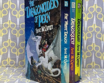 May include: A boxed set of Anne McCaffrey's Dragonriders of Pern books. The box features a fantasy illustration of a dragon and rider. The set includes "The White Dragon", "Dragonquest", and "Dragonflight". The books are in a box with the title "The Dragonriders of Pern".