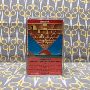 Może przedstawiać: Kaseta magnetofonowa w czerwono-biało-niebieskim pudełku. Pudełko zawiera tekst "The Album Of The Soundtrack Of The Film Of Monty Python And The Holy Grail".