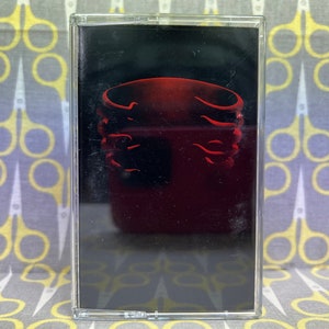 Op de afbeelding: Een zwarte cassetteband met een rode en zwarte afbeelding van een bandlogo. Het logo is een gestileerde afbeelding van een cirkel met drie ringen.