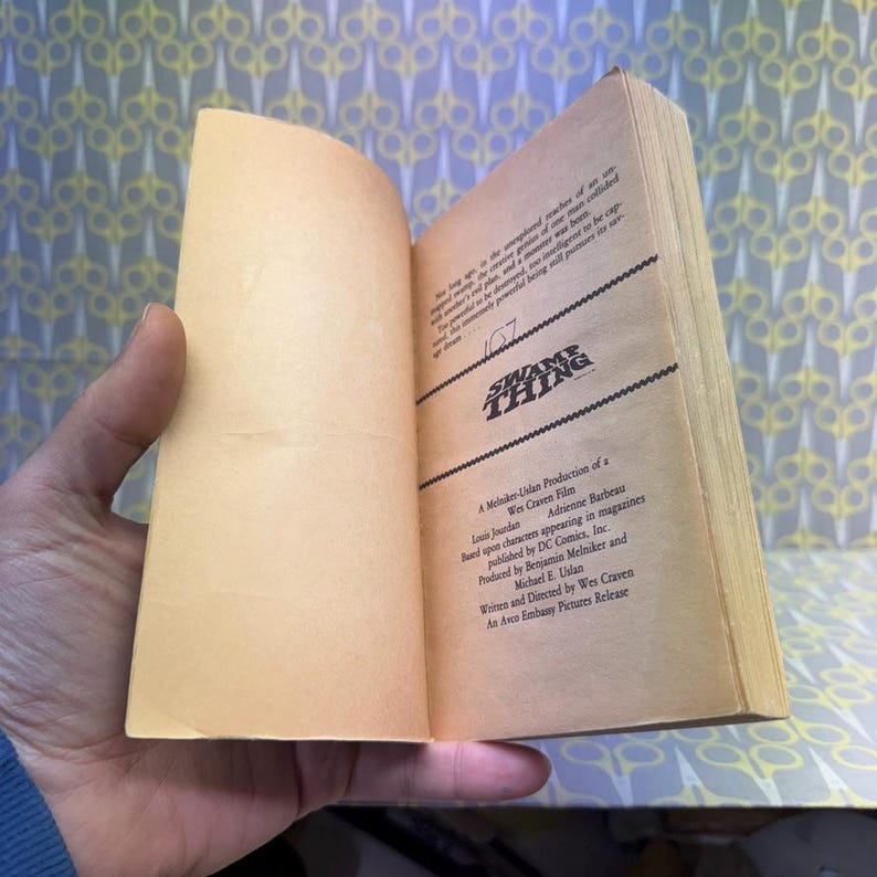 May include: An open book with yellowed pages, featuring the title "SWAMP THING" in bold, black letters. The book's text includes production credits and the names of the cast and crew. The book is held open by a hand.