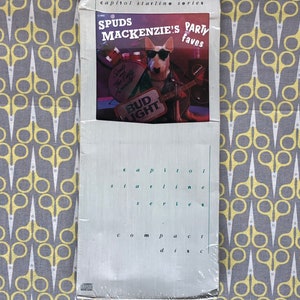 May include: A white compact disc case with a white bull terrier wearing sunglasses and a leather jacket. The case has the text "Capitol Starline Series" and "Spuds Mackenzie's Party Faves" on the front. The bull terrier is holding a guitar and a Bud Light can.
