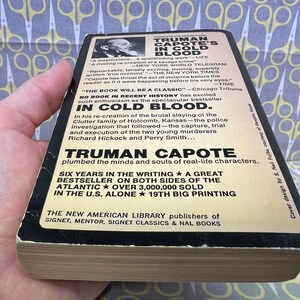 May include: A black and white paperback book cover with the title "TRUMAN CAPOTE'S IN COLD BLOOD". The cover features a black and white photo of Truman Capote and text about the book, including the quote "The book will be a classic." and the fact that it was a bestseller. The book is a re-creation of the brutal slaying of the Clutter family of Holcomb, Kansas.