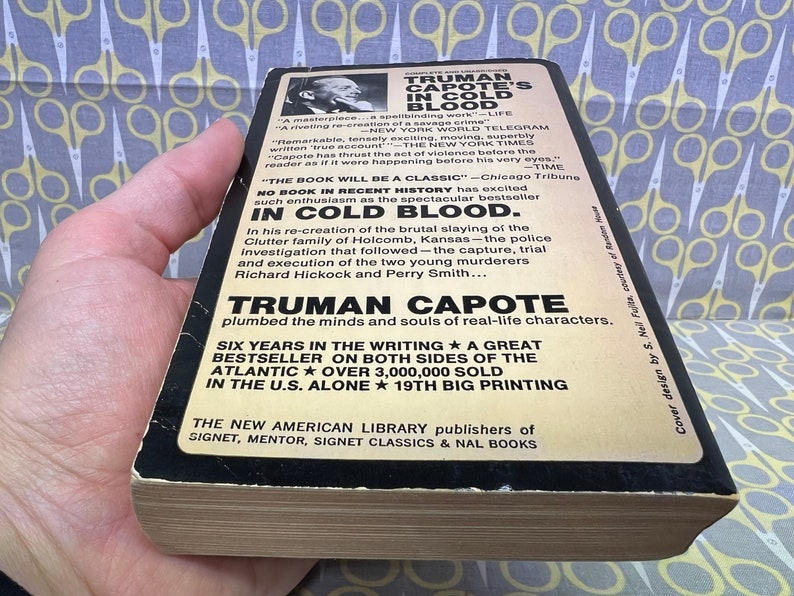 May include: A black and white paperback book cover with the title "TRUMAN CAPOTE'S IN COLD BLOOD". The cover features a black and white photo of Truman Capote and text about the book, including the quote "The book will be a classic." and the fact that it was a bestseller. The book is a re-creation of the brutal slaying of the Clutter family of Holcomb, Kansas.