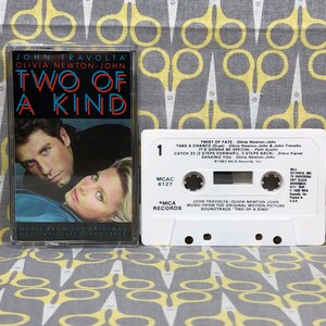 May include: A white cassette tape with the title "Two of a Kind" featuring John Travolta and Olivia Newton-John. The cassette tape is from the original motion picture soundtrack.
