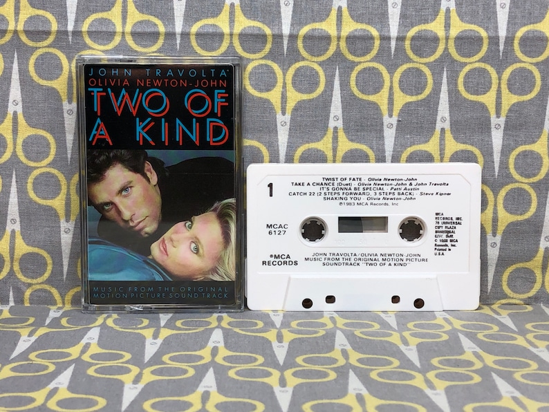 May include: A white cassette tape with the title "Two of a Kind" featuring John Travolta and Olivia Newton-John. The cassette tape is from the original motion picture soundtrack.