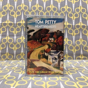 Puede incluir: Una cinta de casete vintage con una imagen pintada de un camino sinuoso que conduce a una casa en un entorno rural. La cinta está etiquetada como "Tom Petty and the Heartbreakers" y "Into the Great Wide Open".