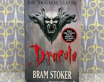 May include: A paperback book titled Dracula by Bram Stoker. The cover features a grey, stone-like gargoyle face with fangs and a red, star-shaped burst containing the text: With 8 pages of photos from the magnificent new motion picture.