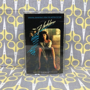 May include: A black cassette tape case with a clear plastic cover. The case features a photo of a woman sitting on a couch, with the title "Flashdance" in blue and white text. The text "Original Soundtrack from the Motion Picture" is printed above the title.