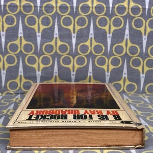May include: A vintage paperback book titled "R is for Rocket" by Ray Bradbury. The book cover features a fiery red and orange illustration of a rocket launch.