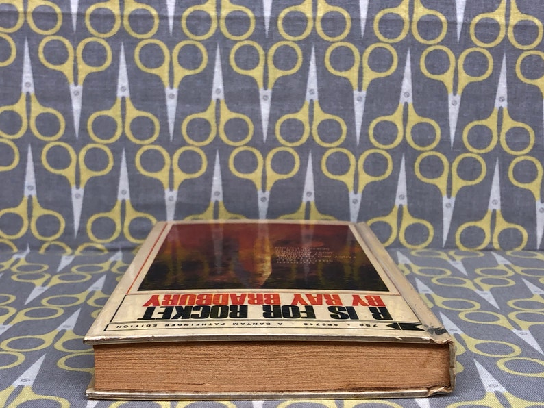 May include: A vintage paperback book titled "R is for Rocket" by Ray Bradbury. The book cover features a fiery red and orange illustration of a rocket launch.