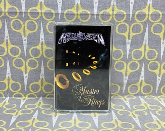 Puede incluir: Una cinta de casete negra con una banda plateada y morada que dice "HELLOWEEN" en letras blancas. La cinta de casete tiene un fondo negro con un diseño de anillos dorados y el texto "Master of the Rings" en letras blancas cursivas.
