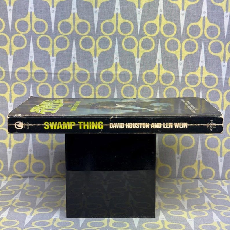 May include: A vintage paperback book titled "Swamp Thing" by David Houston and Len Wein. The cover features a green and black illustration. The book is displayed on a black stand against a patterned background.