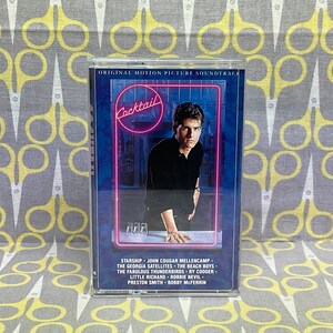 May include: A blue cassette tape case with a photo of Tom Cruise on the front. The case has the title "Cocktail" in pink neon letters. The back of the case lists the songs on the soundtrack, including Starship, John Cougar Mellencamp, The Georgia Satellites, The Beach Boys, The Fabulous Thunderbirds, Ry Cooder, Little Richard, Robbie Nevil, Preston Smith, and Bobby McFerrin.