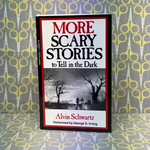 Puede incluir: Una cinta de casete con el t&iacute;tulo "MORE SCARY STORIES to Tell in the Dark" en texto rojo y negro. La portada presenta una ilustraci&oacute;n en blanco y negro de una escena espeluznante con figuras y &aacute;rboles silueteados. Tambi&eacute;n se muestra el nombre del autor, Alvin Schwartz.