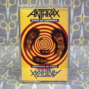 Puede incluir: Una cinta de cassette con una cubierta amarilla. El nombre de la banda "ANTHRAX" está en negro, y el título del álbum "STATE OF EUPHORIA" está en azul. La portada presenta un diseño en espiral con caras gritando en rojo y amarillo.