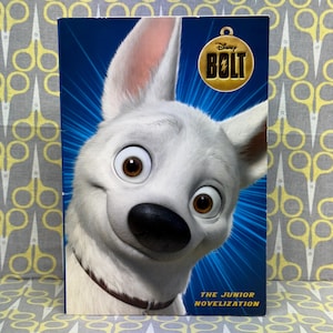 Può includere: La copertina di un libro presenta un cane bianco con occhi marroni e naso nero. Il libro è intitolato "Disney Bolt: La novellizzazione junior".
