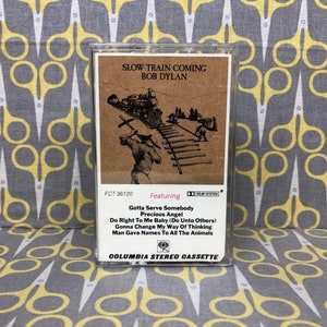 May include: A vintage cassette tape with a brown and white illustration of a train on the front. The cassette tape is labeled "Slow Train Coming" and "Bob Dylan". The text "Featuring" is printed below the image, followed by a list of song titles: "Gotta Serve Somebody", "Precious Angel", "Do Right To Me Baby (Do Unto Others)", "Gonna Change My Way Of Thinking", and "Man Gave Names To All The Animals". The cassette tape is labeled "Columbia Stereo Cassette".