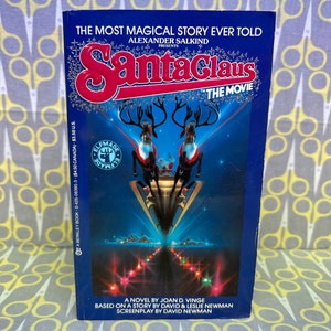 May include: A blue book cover with the title "Santa Claus: The Movie" in red and silver lettering. The cover features a scene of Santa Claus flying in his sleigh pulled by reindeer over a city skyline. The book is based on a novel by Joan D. Vinge and a story by David and Leslie Newman.