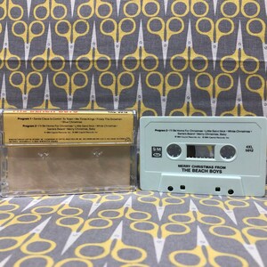 May include: A white cassette tape with the text "Merry Christmas from The Beach Boys" and "4XL 5012" printed on it. The cassette tape is in a clear plastic case with a gold label that lists the songs on the tape.