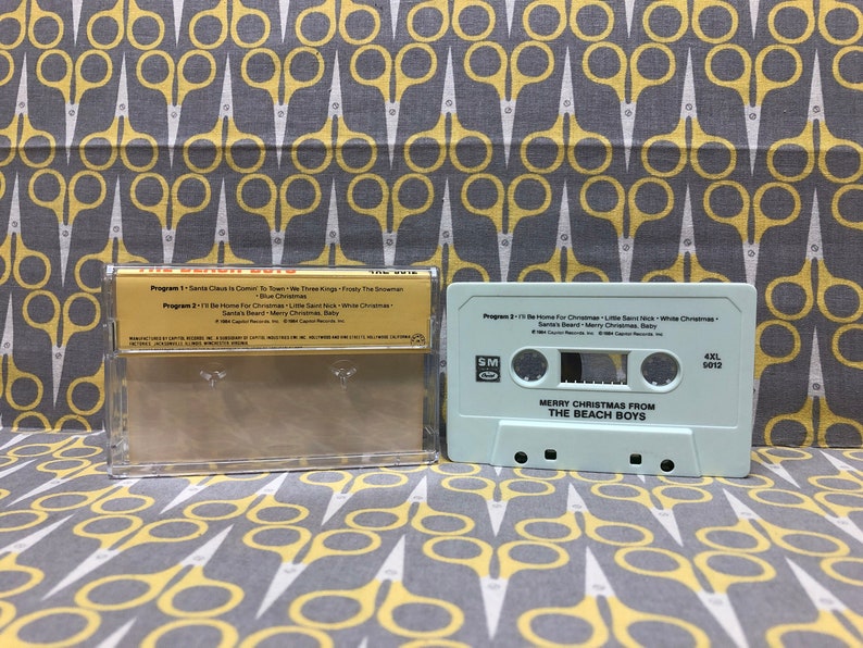 May include: A white cassette tape with the text "Merry Christmas from The Beach Boys" and "4XL 5012" printed on it. The cassette tape is in a clear plastic case with a gold label that lists the songs on the tape.
