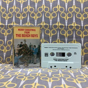 May include: A vintage cassette tape with a light blue case. The tape is labeled "Merry Christmas from The Beach Boys". The front cover of the cassette tape features a photo of The Beach Boys standing around a Christmas tree.
