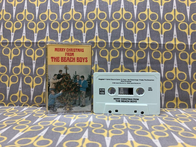 May include: A vintage cassette tape with a light blue case. The tape is labeled "Merry Christmas from The Beach Boys". The front cover of the cassette tape features a photo of The Beach Boys standing around a Christmas tree.