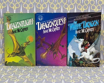 Könnte beinhalten: Drei Taschenbücher aus der Reihe Dragonriders of Pern von Anne McCaffrey. Die Titel lauten Dragonflight, Dragonquest und The White Dragon. Jedes Cover zeigt farbenfrohe Fantasy-Illustrationen von Drachen und ihren Reitern.
