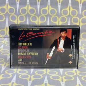May include: A black cassette tape case with a photo of a man holding a guitar. The case has the title "La Bamba" and the text "Original Motion Picture Soundtrack" on the front. The text "Performed by Los Lobos, Bo Diddley, Howard Huntsberry, Brian Setzer and Marshall Crenshaw" is also on the front of the case.