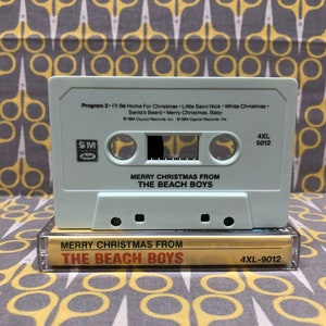 May include: A white cassette tape with the title "Merry Christmas From The Beach Boys" and the song list "I'll Be Home For Christmas", "Little Saint Nick", "White Christmas", "Santa's Beard", and "Merry Christmas, Baby". The cassette tape is in a gold cassette case with the same title.