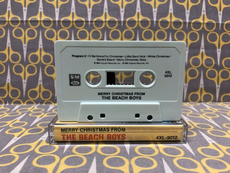May include: A white cassette tape with the title "Merry Christmas From The Beach Boys" and the song list "I'll Be Home For Christmas", "Little Saint Nick", "White Christmas", "Santa's Beard", and "Merry Christmas, Baby". The cassette tape is in a gold cassette case with the same title.