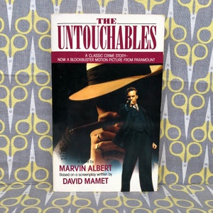Puede incluir: Una portada de libro de bolsillo con una foto en blanco y negro de un hombre con traje sosteniendo un arma. El título del libro es "Los Intocables" y el autor es Marvin Albert. El libro está basado en un guion escrito por David Mamet.