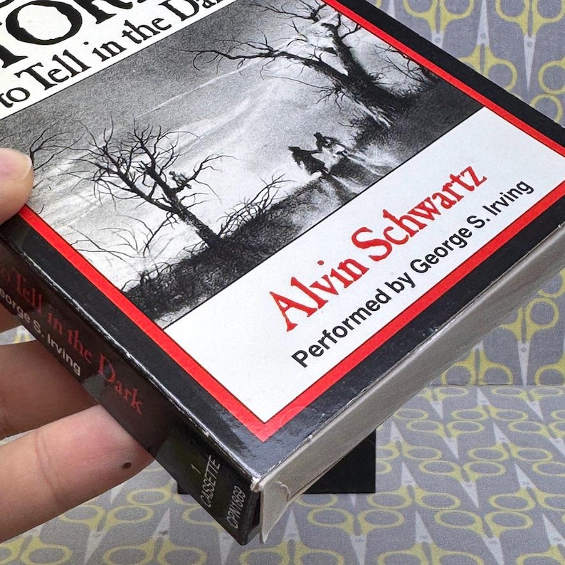 Puede incluir: Caja de cinta de cassette para "Historias de miedo para contar en la oscuridad" de Alvin Schwartz. La caja presenta una ilustraci&oacute;n en blanco y negro de una escena espeluznante con &aacute;rboles desnudos y figuras. El t&iacute;tulo y el nombre del autor son en rojo.