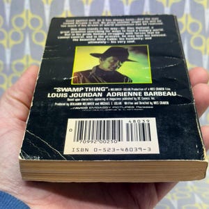 May include: A vintage paperback book titled "Swamp Thing" with a black cover featuring a man in a hat. The book's spine is visible, showing the title and author's names. The book is held in a person's hand.
