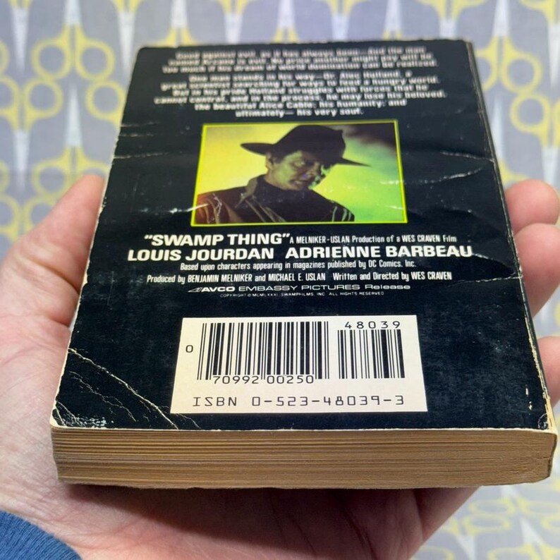 May include: A vintage paperback book titled "Swamp Thing" with a black cover featuring a man in a hat. The book's spine is visible, showing the title and author's names. The book is held in a person's hand.