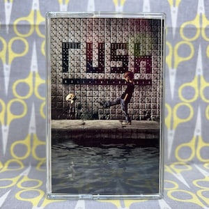 May include: A cassette tape with a black and white image of a young person kicking a skull against a wall covered in dice. The wall has the word "DICE" written on it.