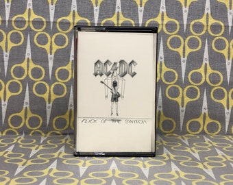May include: A black cassette tape case with a white label. The label has a black and white illustration of a person holding a guitar. The text "AC/DC" is printed in black above the illustration and "FLICK OF THE SWITCH" is printed in black below the illustration.