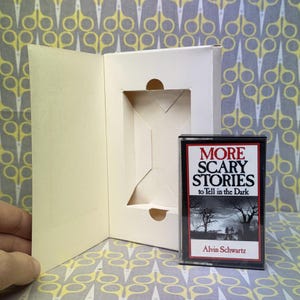 Puede incluir: Una cinta de cassette vintage de "M&aacute;s historias de miedo para contar en la oscuridad" de Alvin Schwartz, con una portada blanca y roja. La cinta est&aacute; en una caja de cart&oacute;n blanca con un recorte.
