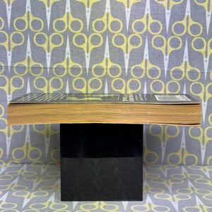 May include: A book with yellowed pages and a black cover, resting on a black stand. The background features a repeating pattern of yellow and white scissors on a gray fabric.
