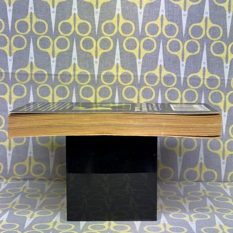 May include: A book with yellowed pages and a black cover, resting on a black stand. The background features a repeating pattern of yellow and white scissors on a gray fabric.