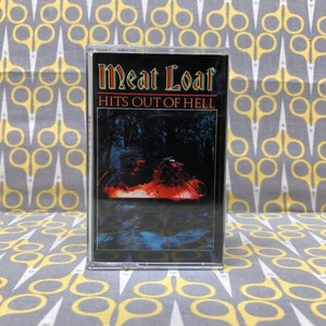 May include: A cassette tape with a black and white image of a fiery scene in a forest. The text on the cassette tape reads "Meat Loaf Hits Out of Hell."
