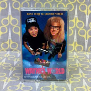 May include: A cassette tape for the movie "Wayne's World" featuring a blue background with the film's stars. The tape case has the movie title in pink and the text "MUSIC FROM THE MOTION PICTURE".