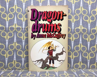 May include: A book cover with a yellow, orange, and red title "Dragon-drums" by Anne McCaffrey. The cover features an illustration of a young person holding a small, golden dragon.