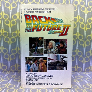Könnte beinhalten: Ein Taschenbuchcover für den Roman "Zurück in die Zukunft II", basierend auf dem gleichnamigen Film. Das Cover zeigt eine Collage von Bildern aus dem Film, darunter das DeLorean-Zeitfahrzeug, Marty McFly und Doc Brown.