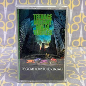 May include: A Teenage Mutant Ninja Turtles cassette tape. The cover features the title in green, the turtles' faces, and a cityscape. The text "THE ORIGINAL MOTION PICTURE SOUNDTRACK" is at the bottom.