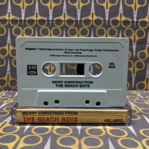 May include: A light blue cassette tape with the title "Merry Christmas From The Beach Boys" and the song list "Santa Claus Is Comin' To Town, We Three Kings, Frosty The Snowman, Blue Christmas". The cassette tape has a white label with black text and a black and white label with white text.
