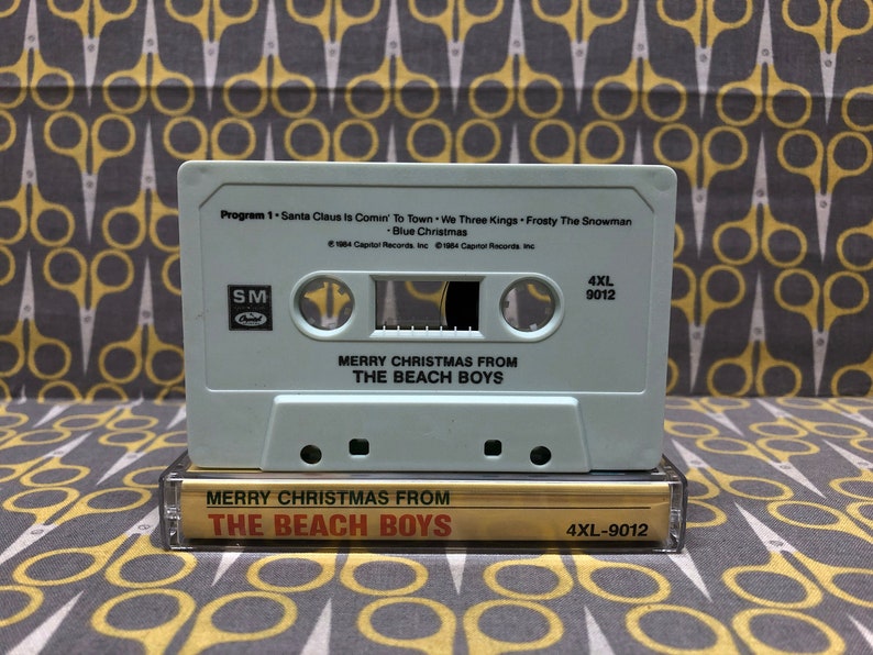 May include: A light blue cassette tape with the title "Merry Christmas From The Beach Boys" and the song list "Santa Claus Is Comin' To Town, We Three Kings, Frosty The Snowman, Blue Christmas". The cassette tape has a white label with black text and a black and white label with white text.